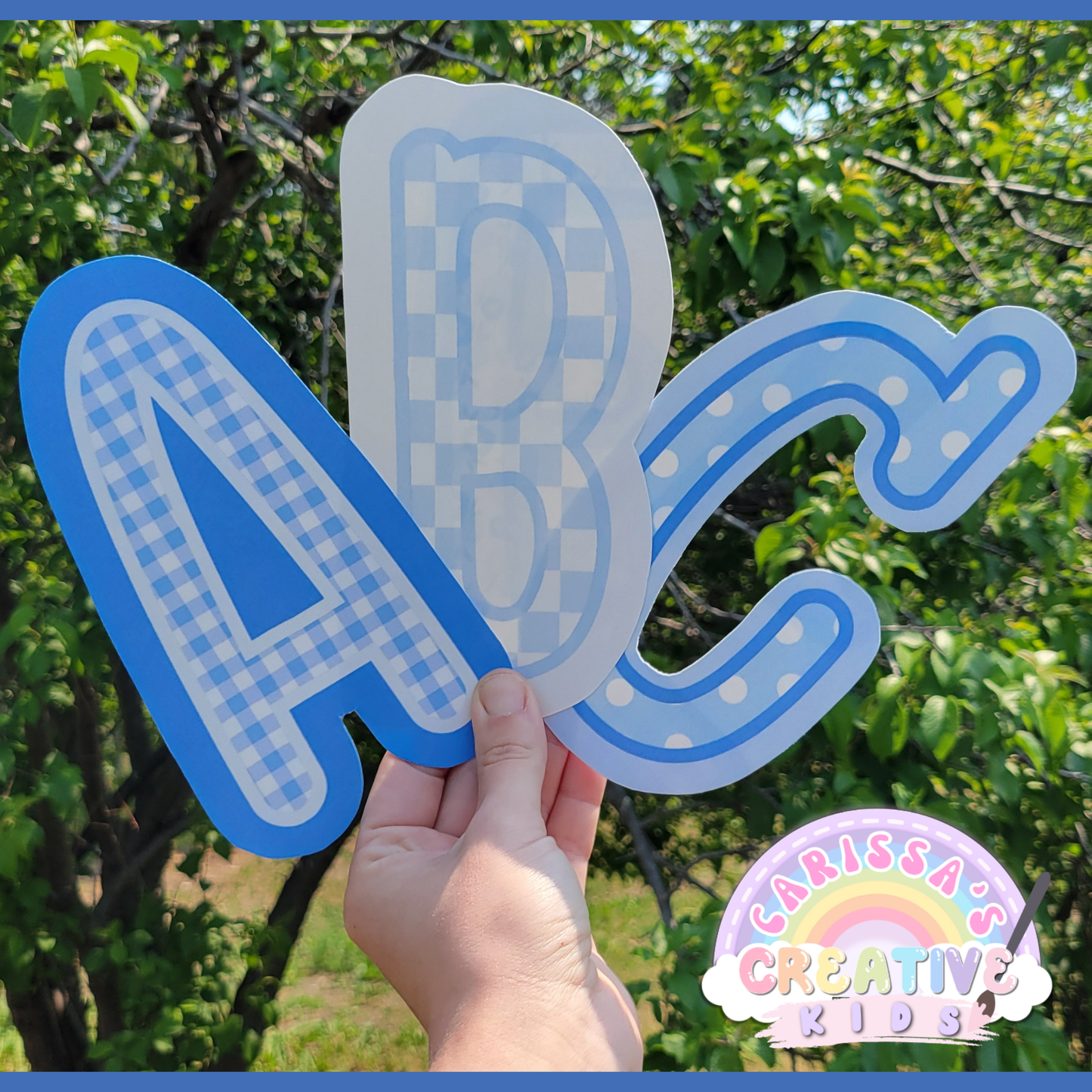 Hand holding cut-out bulletin board letters A, B, and C from the printable Silly Goose collection. Letters feature gingham, polka dot, and checkered blue patterns. Shown outdoors to highlight size and print quality, perfect for classroom door or board decor.